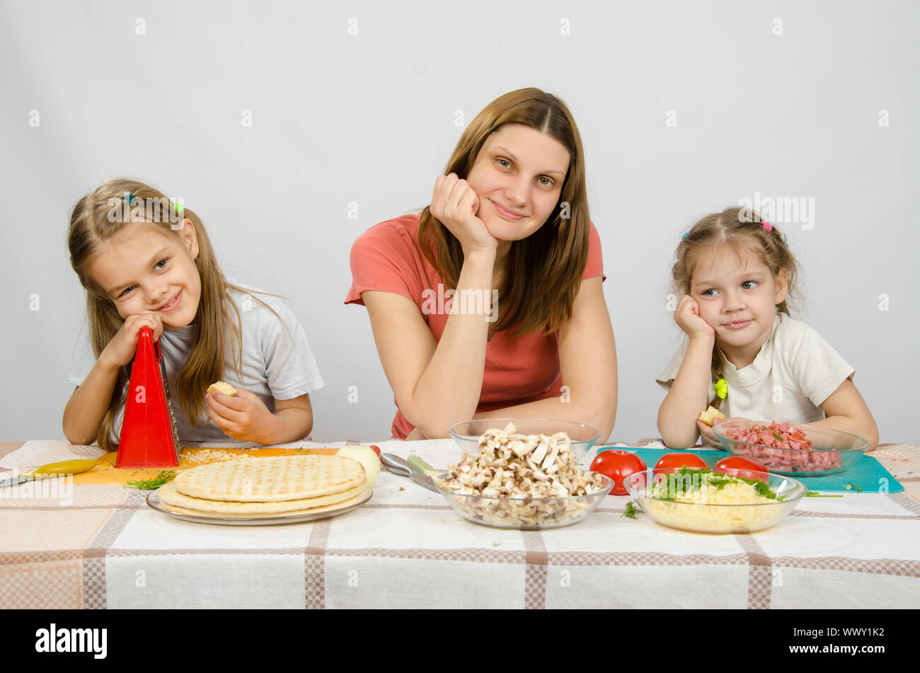 Mère satisfait avec deux filles assis reposant sa tête dans ses mains à la table avec les produits pour pizza Banque D'Images