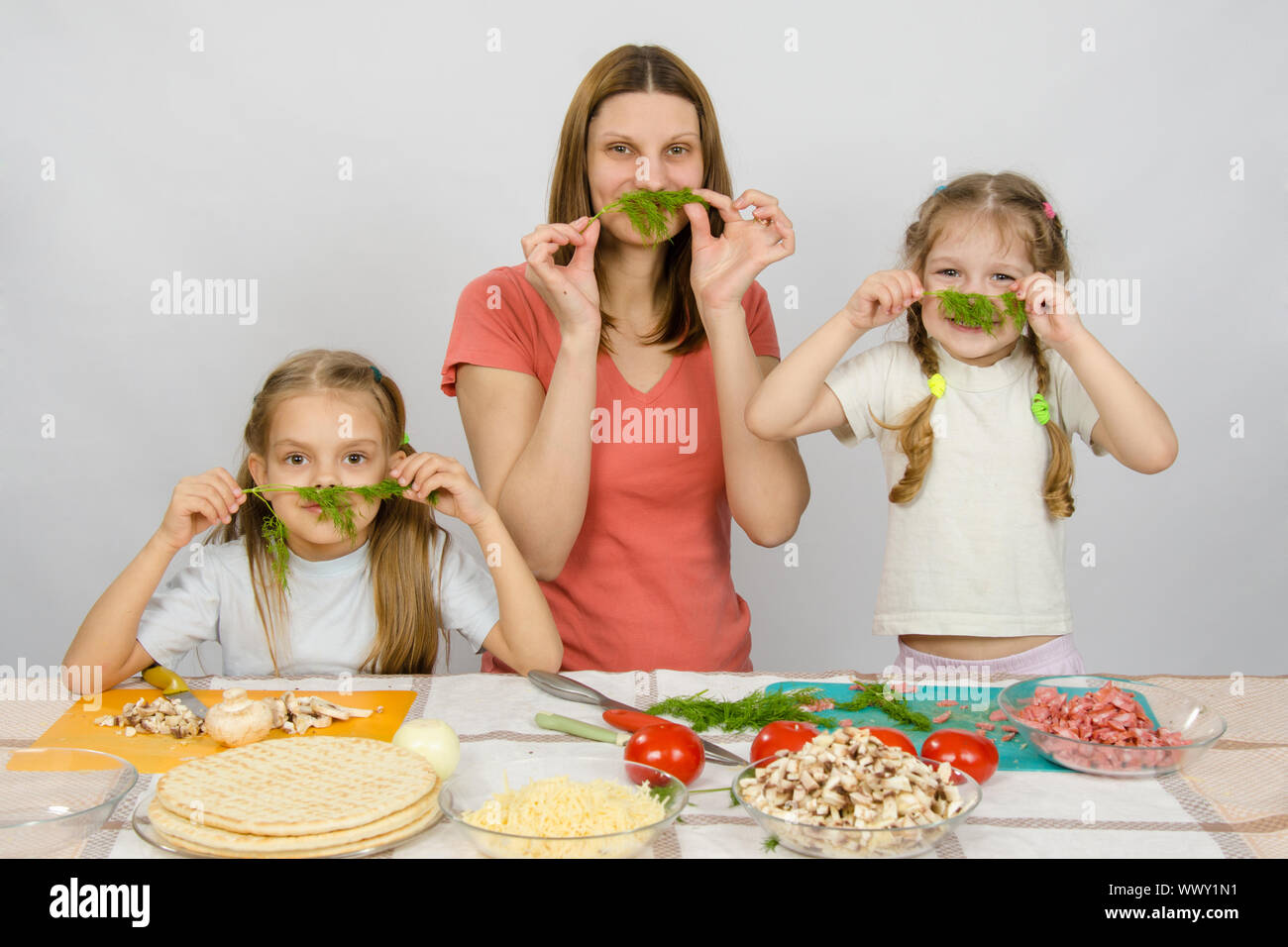 Jeune femme au foyer avec deux filles s'amusant tenant un brin de persil comme une moustache à la table de cuisine lorsqu'il partage la cuisson Banque D'Images