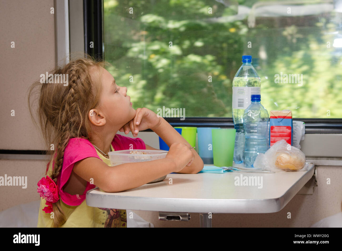 Une fille de six ans dans un train assis à une table avec de la nourriture sur une place inférieure dans le compartiment de deuxième classe de la voiture et a l'air e Banque D'Images