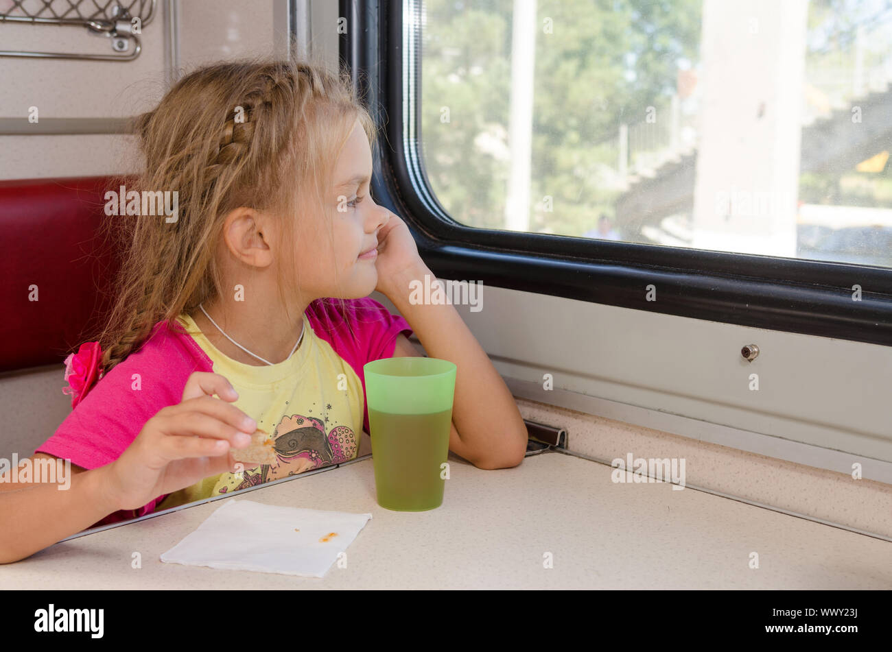 Six ans fille assise dans le train à la table sur l'extérieur du chariot de deuxième classe et avec enthousiasme par la fenêtre Banque D'Images