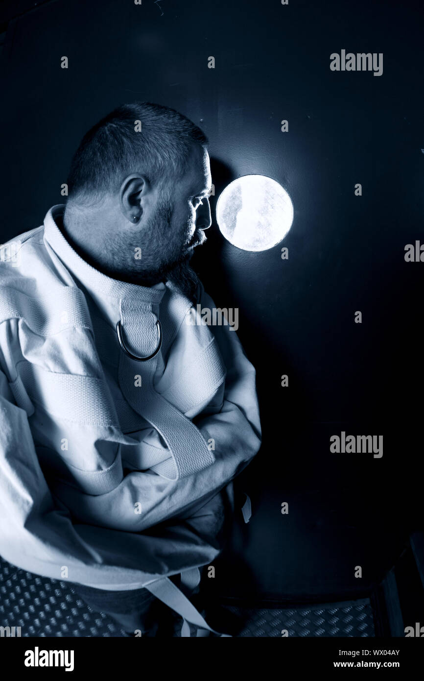 Photo d'un aliéné dans la quarantaine portant une camisole debout dans une cellule d'un asile avec la lumière du couloir en streaming. Banque D'Images