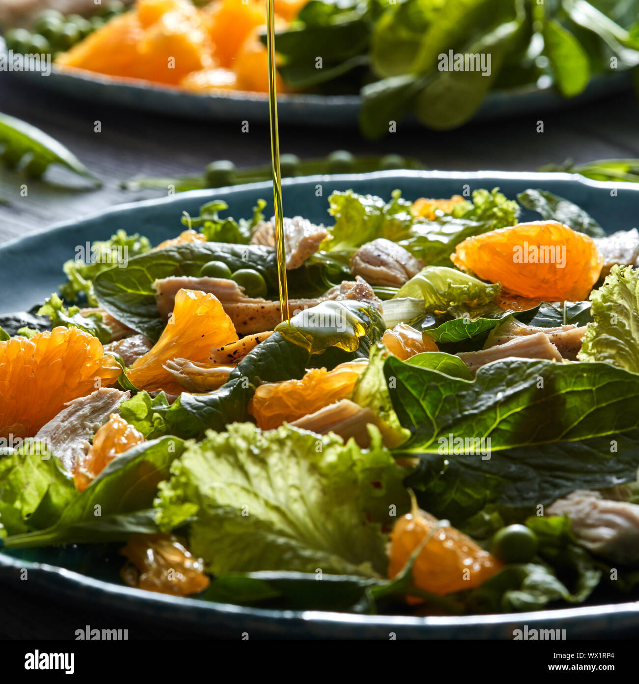 Un filet d'huile d'olive se déverse dans la salade préparée avec des légumes verts, morceaux de poulet, les tranches d'orange dans un bleu ceram Banque D'Images