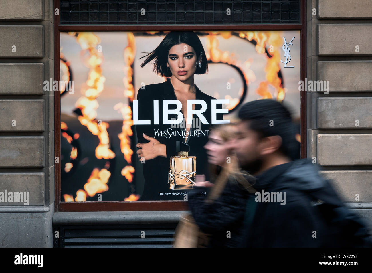 Couple (blurred motion) balade par un advetisement pour le parfum Libre par Yves Saint Laurent YSL dans la fenêtre de magasin Jenners, Édimbourg. Banque D'Images