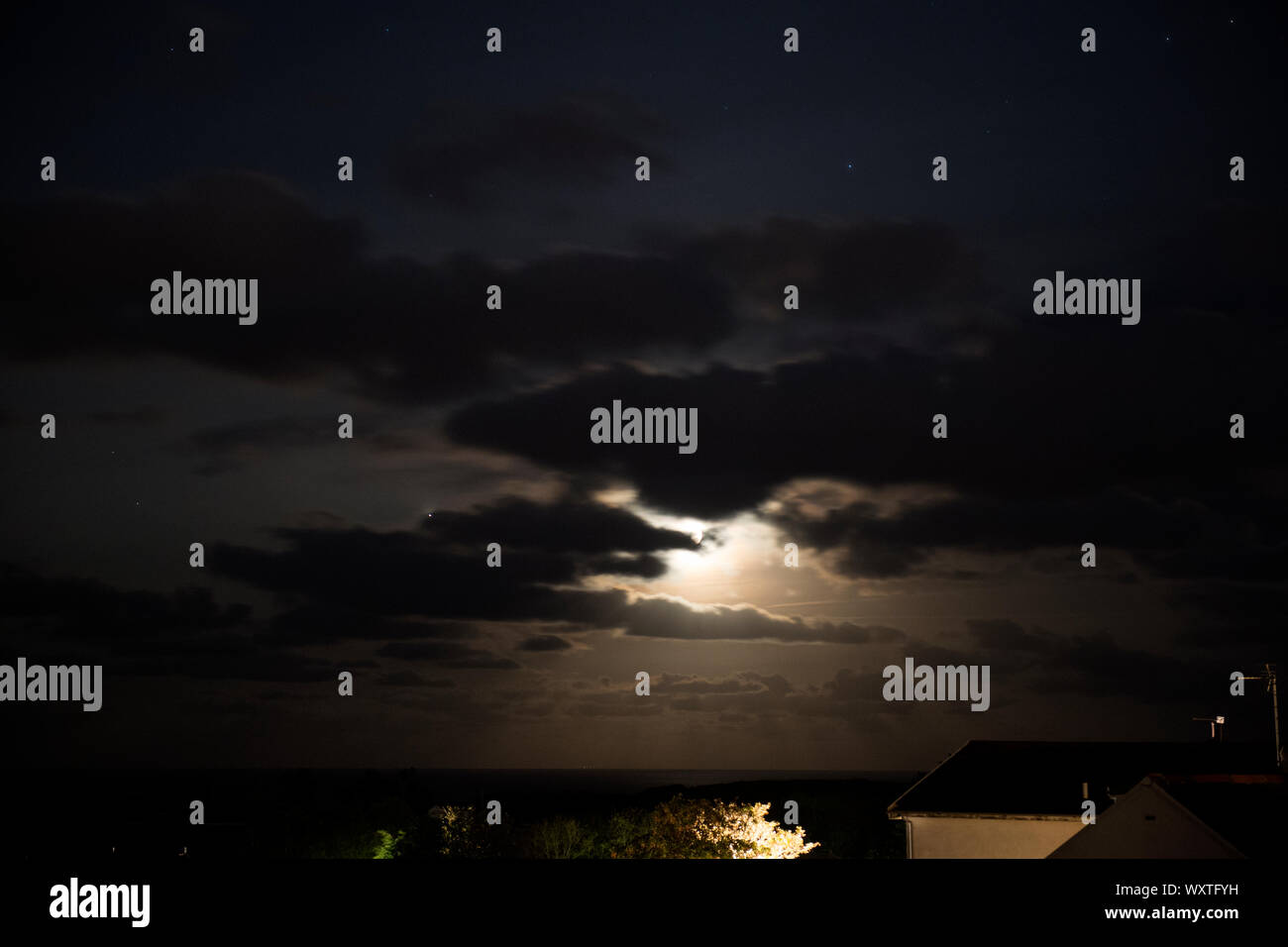 Lune brille à travers les nuages sur une mer d'étoiles dans le ciel et les toits visibles. Banque D'Images