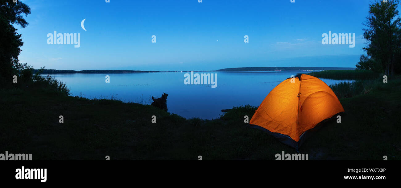 Tourisme Orange tente sur la rive du lac au crépuscule. La lune dans le ciel. Le concept du voyage, de la liberté et de la vie privée Banque D'Images