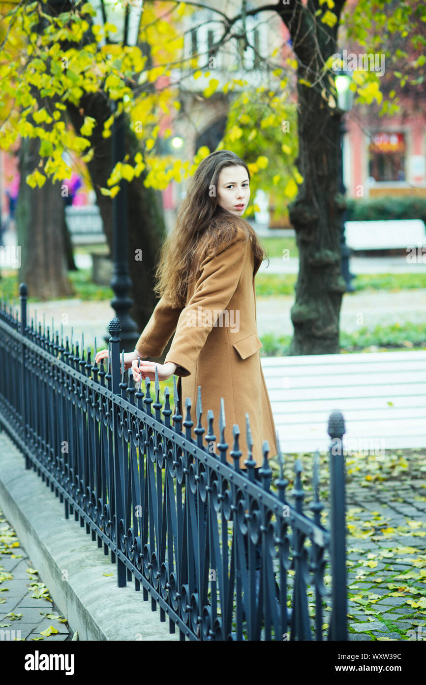 Jeune femme en manteau camel promenades dans le parc de l'automne. Fille  avec de longs cheveux brun près de clôture métallique avec l'arrière-plan  flou, sur le parc. Petite amie en attente de