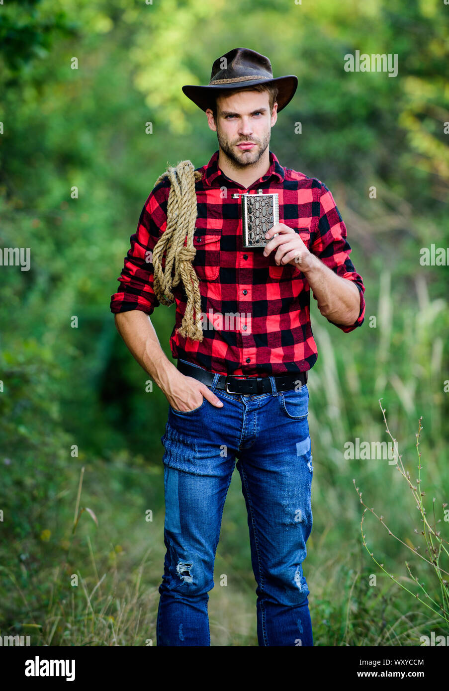 Ranch Cowboy travailleur. Bourbon whiskey. La culture occidentale. Man wearing hat tenir la corde et ballon. Outil Lasso American cowboy. Cowboy brutal de boire de l'alcool. L'homme beau cowboy fond nature. Banque D'Images