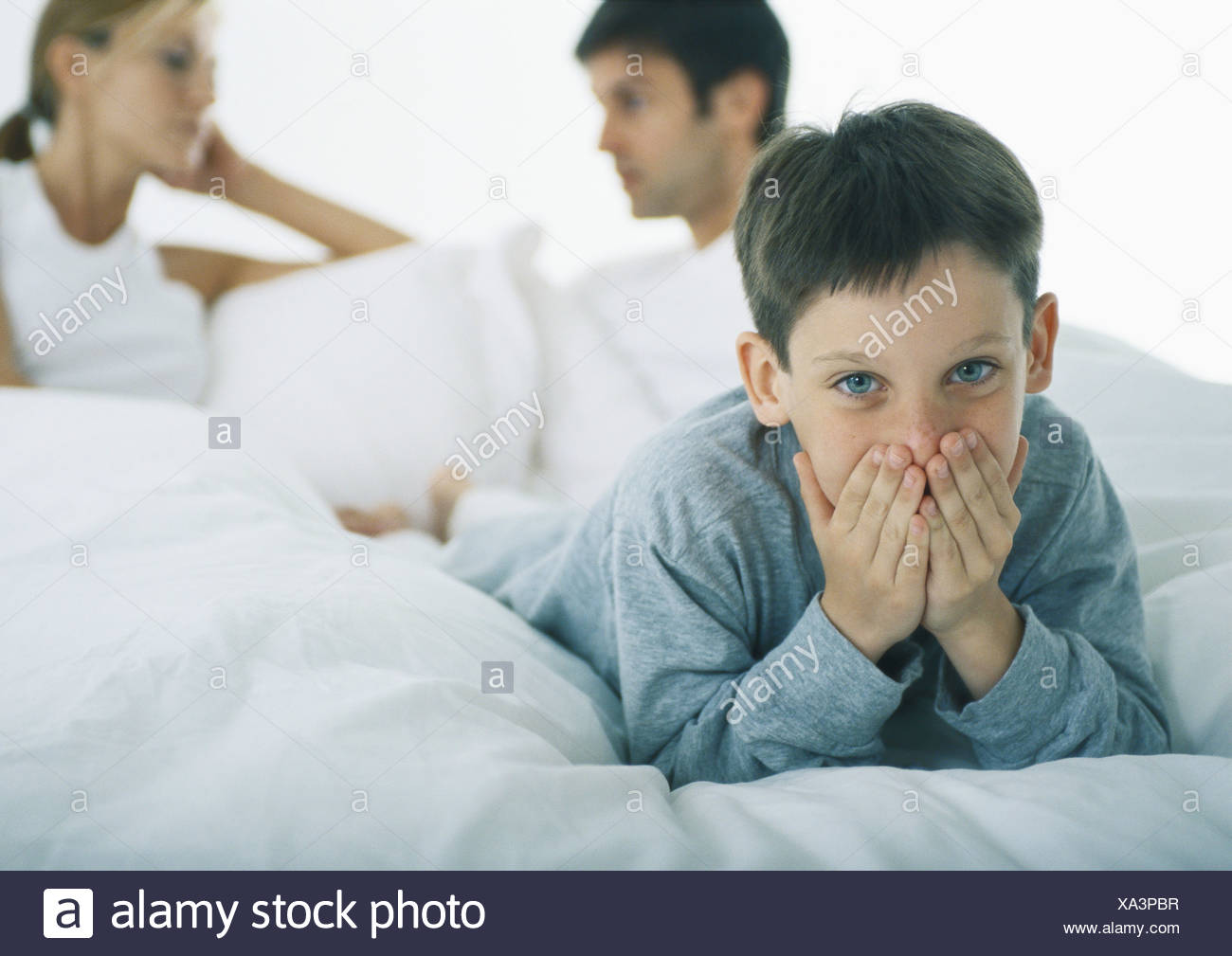 Boy lying on stomach in bed couvrant la bouche avec les mains, les parents de discuter en arrière-plan Banque D'Images