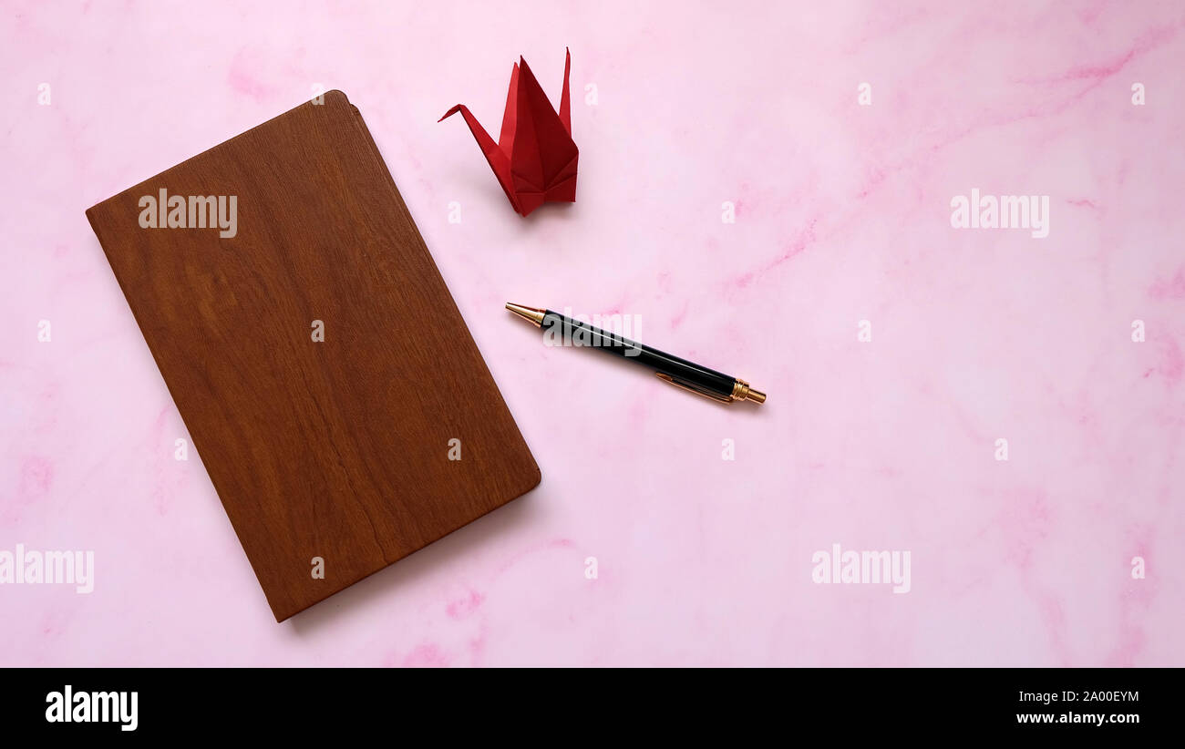 Un notebook con copertura in legno, con una penna a sfera e una carta rossa gru accanto ad esso. Marmo rosa sfondo. Foto Stock