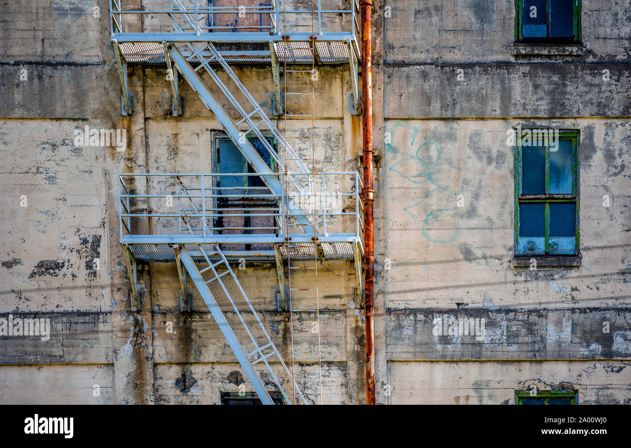 Peeling intonacata parete di un vecchio industriale abbandonato edificio a più piani con finestre rotte e un metallo verniciato scala antincendio e tubi arrugginito Foto Stock