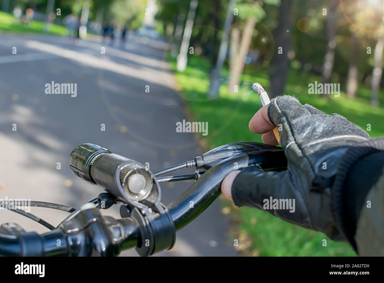 Sigaretta nella mano di un ciclista per fumatori che corre in bici sullo sfondo del percorso asfaltata del parco della città Foto Stock