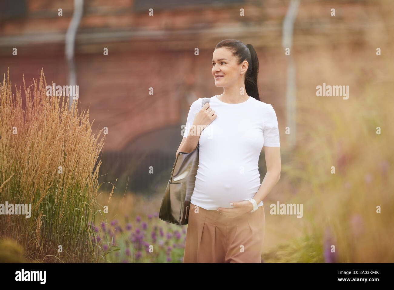 Giovane donna incinta con capelli scuri di indossare un abbigliamento informale a piedi in città e sorridente Foto Stock