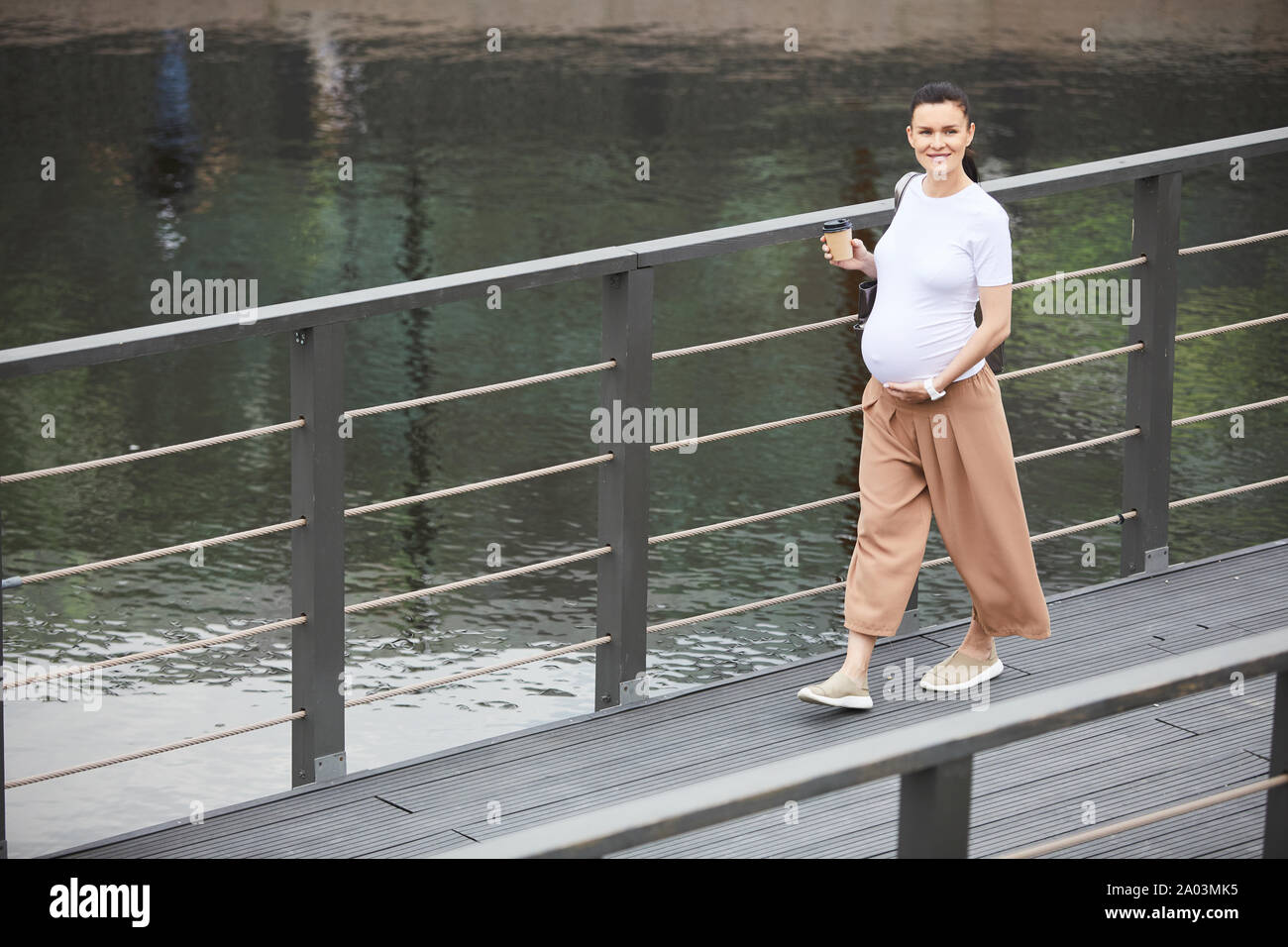 Ritratto di giovane donna incinta sorridente alla videocamera mentre si beve il caffè e camminare lungo il ponte all'aperto Foto Stock