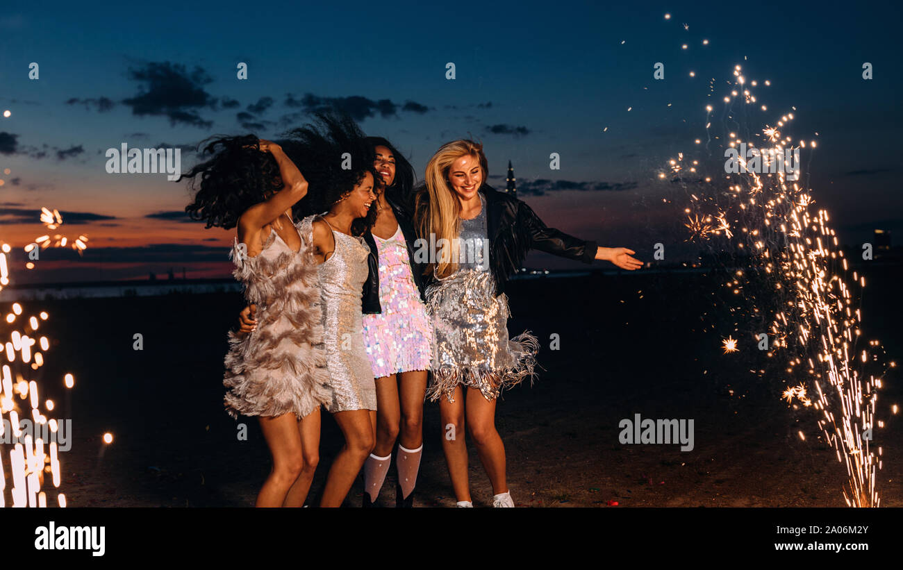Gruppo di amiche felice danza tra i fuochi d'artificio di notte. Giovani donne eleganti celebrando al tramonto. Foto Stock