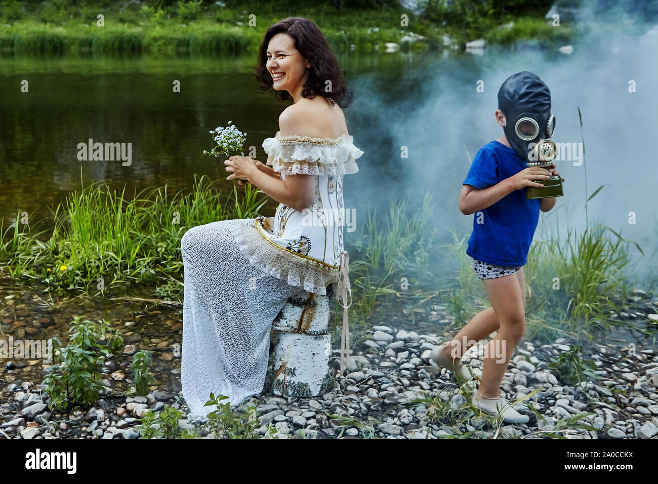 Una bella donna adulta ride, seduto sulla riva del fiume con un mazzo di fiori selvatici nelle sue mani, indossando un matrimonio o un abito da sera e un bambino mi Foto Stock