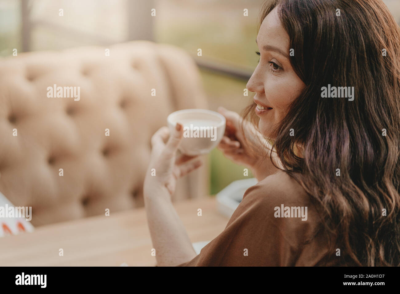 Affascinante brunette donna con lunghi capelli ricci seduta alla finestra nel cafe con la tazza di caffè nelle mani Foto Stock
