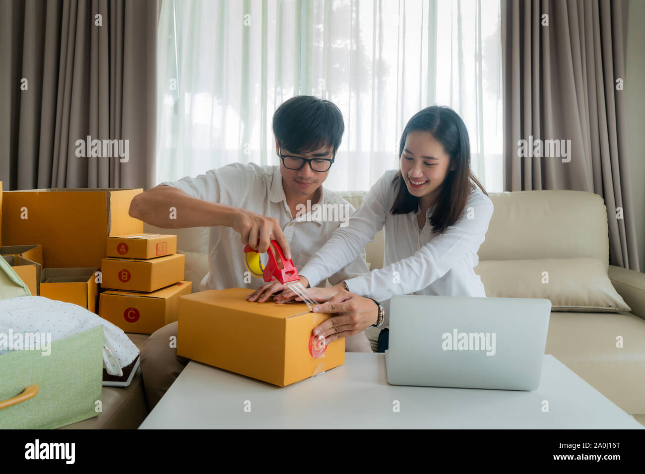 Asian coppia giovane sono la vendita on-line tramite un computer e contribuendo a pack l'ordine del cliente nelle caselle di testo per inviare la posta nel soggiorno di casa. Piccolo Foto Stock