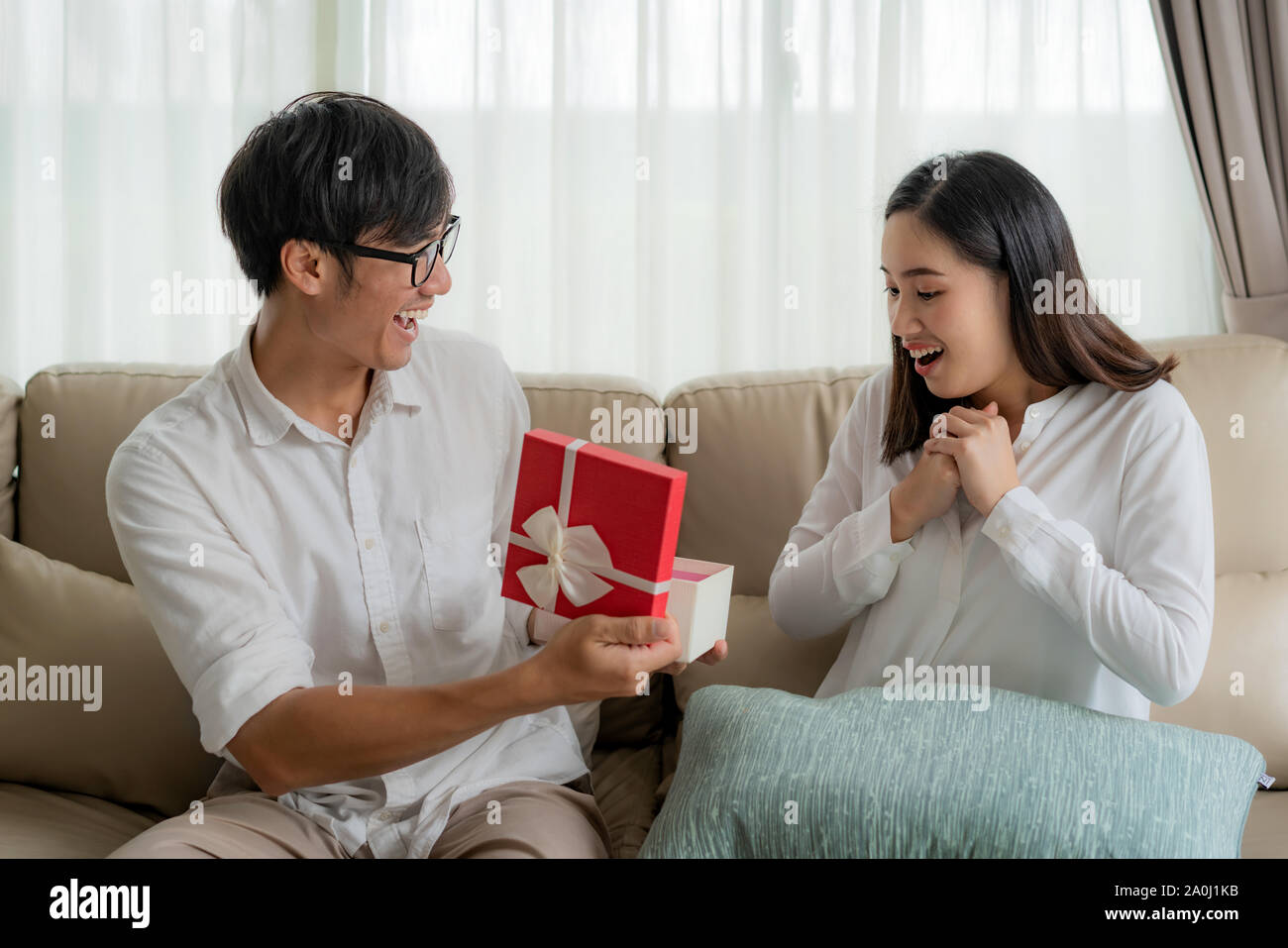 Uomo asiatico dare una donna una confezione regalo rossa. Lei guarda il dono nella scatola e sorpresa per il nostro anniversario di matrimonio o la sua festa di compleanno nel salotto di casa Foto Stock