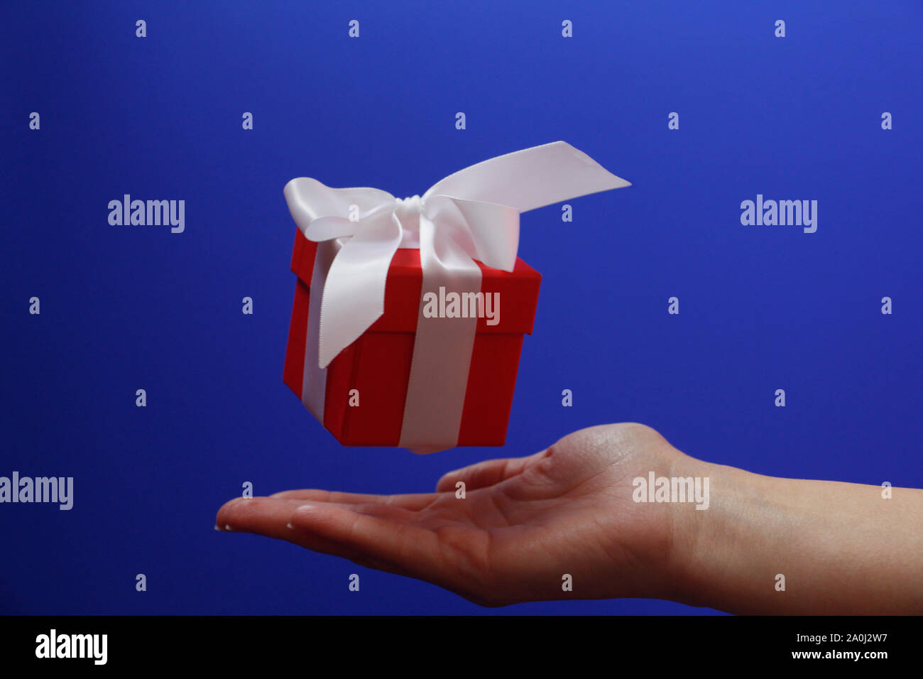 Confezione regalo rossa con un fiocco bianco su sfondo blu. La scatola è chiusa da un coperchio. Dono vola in aria. Al di sotto del dono è una femmina di mano. Concetto. Giv Foto Stock