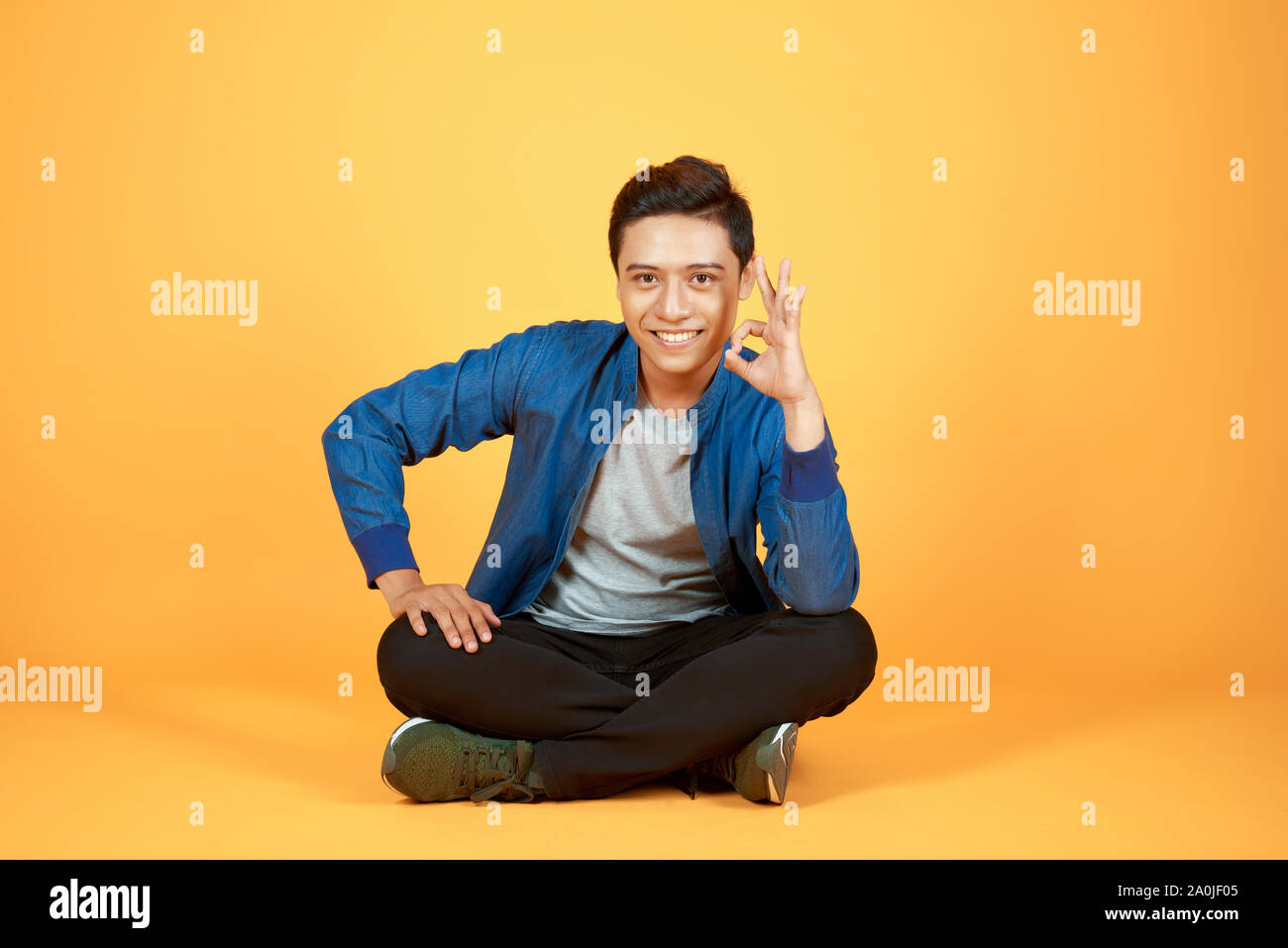 Giovani asiatici casual uomo seduto con le gambe incrociate e tenendo la sua testa mentre sorridente alla telecamera. isolato sul colore di sfondo arancione Foto Stock