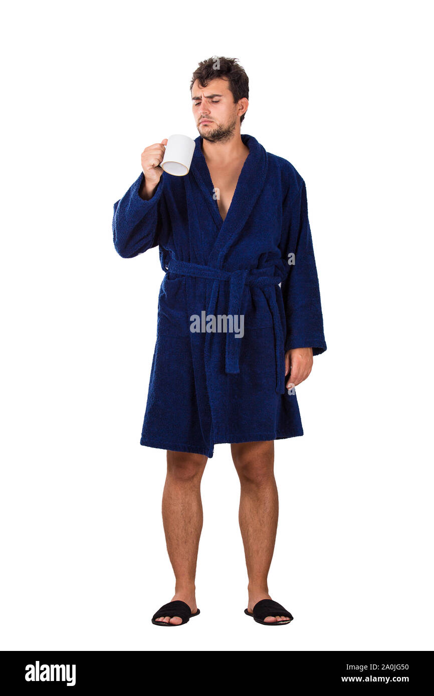 A piena lunghezza Ritratto di sleepy giovane disordinati capelli, indossa accappatoio blu tenendo una tazza di tè in mano cercando confuso isolato su bianco backgrou Foto Stock