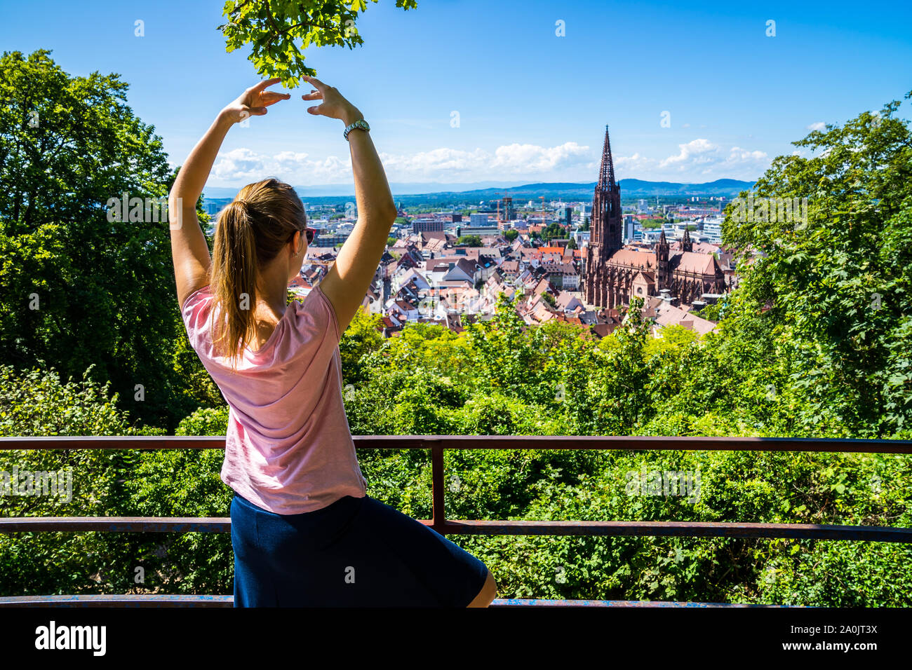 Germania, donna bionda con la coda di cavallo facendo yoga meditazione sopra città Freiburg im Breisgau e skyline con minster cathedral in estate circondata da g Foto Stock