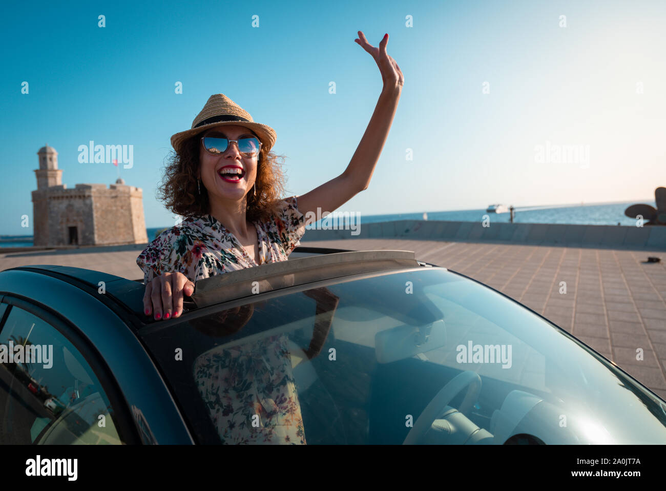 Donna sorridente con cappello di paglia e occhiali da sole godendo di estate in una vettura convertibile. mano saluto. Il concetto di strada viaggi e avventura. equitazione con Foto Stock
