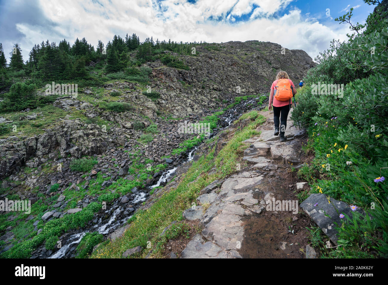 La bionda ragazza adolescente con zaino arancione passeggiate lungo un torrente di montagna su un sentiero roccioso in San Juan Mountains su un nuvoloso giorno di estate, Weminuche selvatica Foto Stock