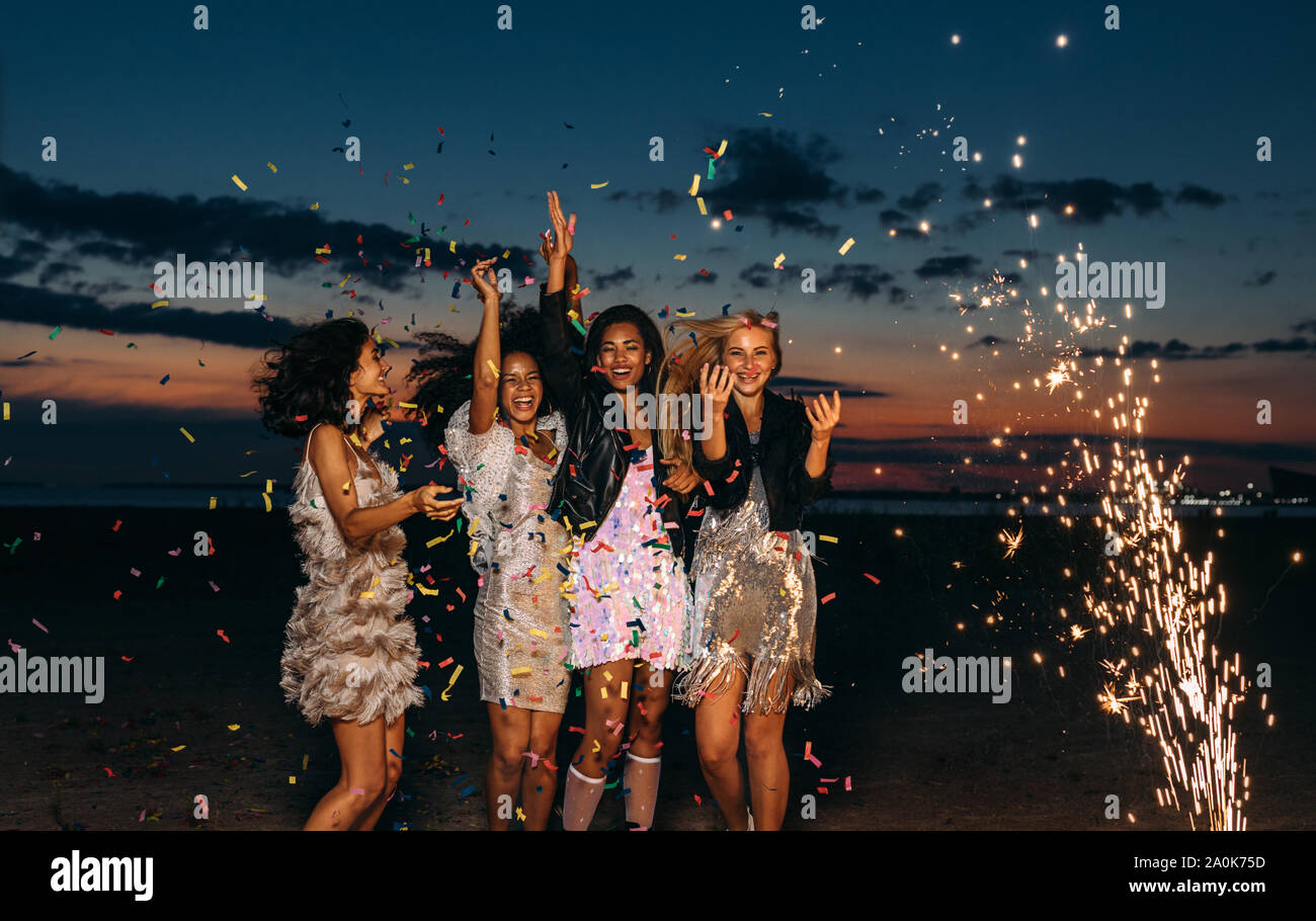 Happy amici celebrando all'aperto al tramonto. Quattro eleganti donne gettando coriandoli all'aperto nei pressi di fuochi d'artificio. Foto Stock