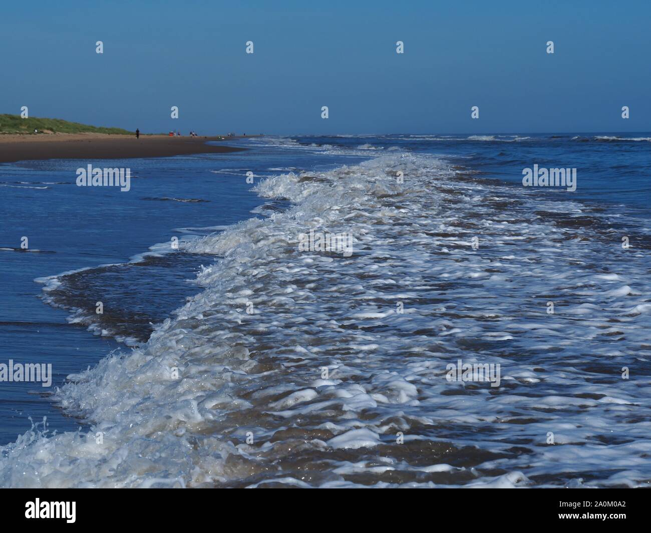 Le onde che si infrangono in avvicinamento alla spiaggia sabbiosa di Mablethorpe, Lincolnshire, Inghilterra, con un cielo blu chiaro Foto Stock