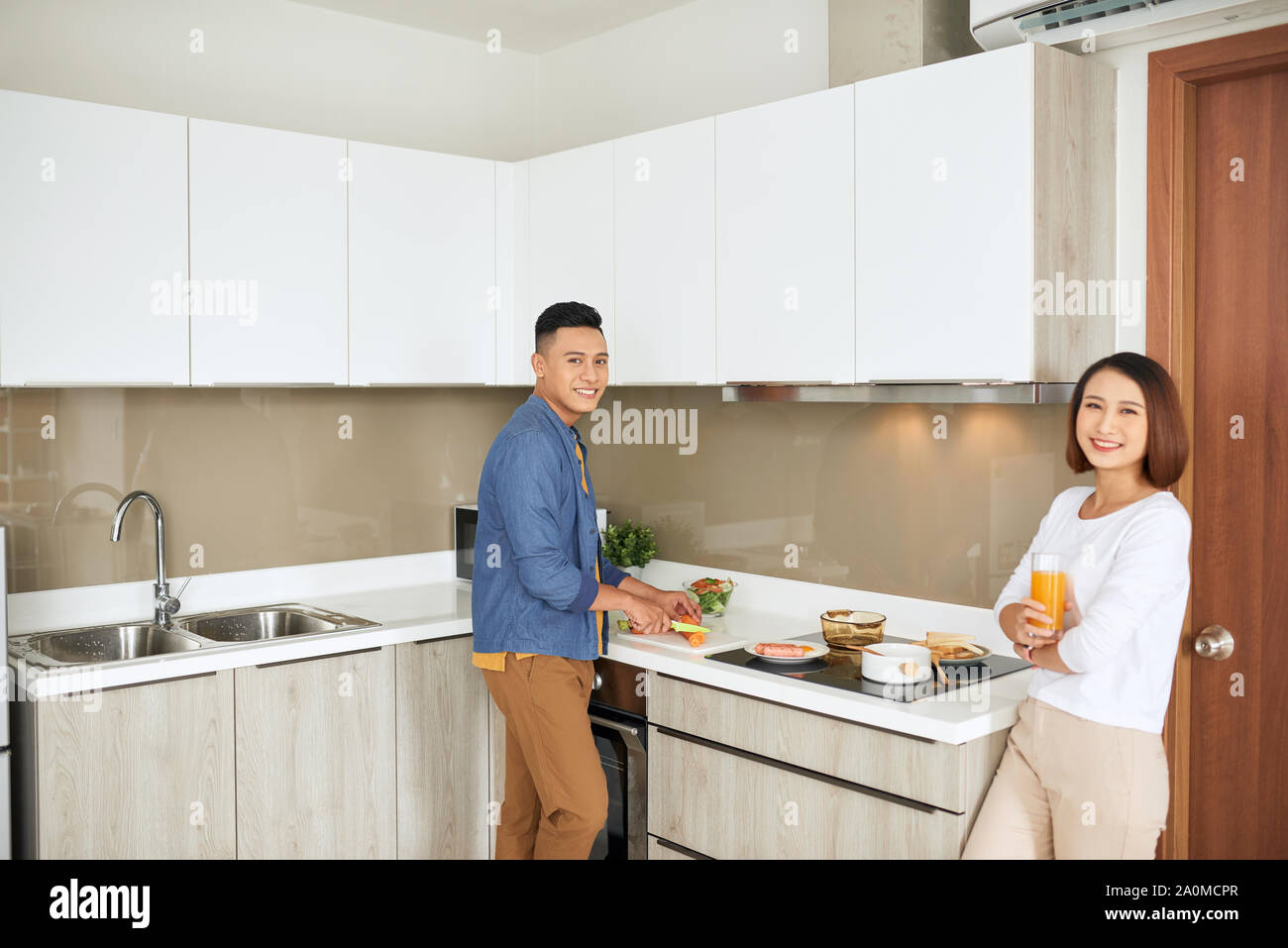 Coppia giovane avente la colazione, donna che mantiene il succo d'arancia durante l uomo di cottura degli alimenti cucina moderno appartamento interno Foto Stock