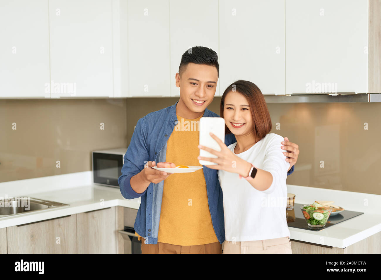 Romantico giovane coppia asiatica cucina insieme e utilizzando il telefono cellulare per selfie in bianco la cucina moderna, smart home concetto Foto Stock