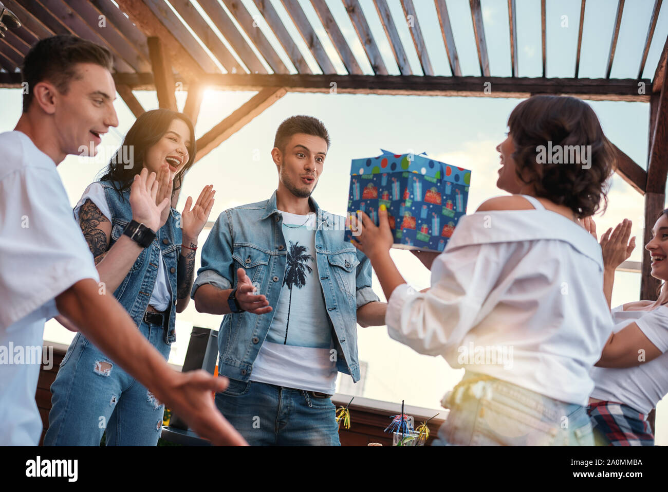 Sorpresa giovane donna è in possesso di una scatola regalo per il suo amico mentre si sta in piedi sul tetto intorno gli amici. Celebrando. Parte. Concetto di compleanno Foto Stock