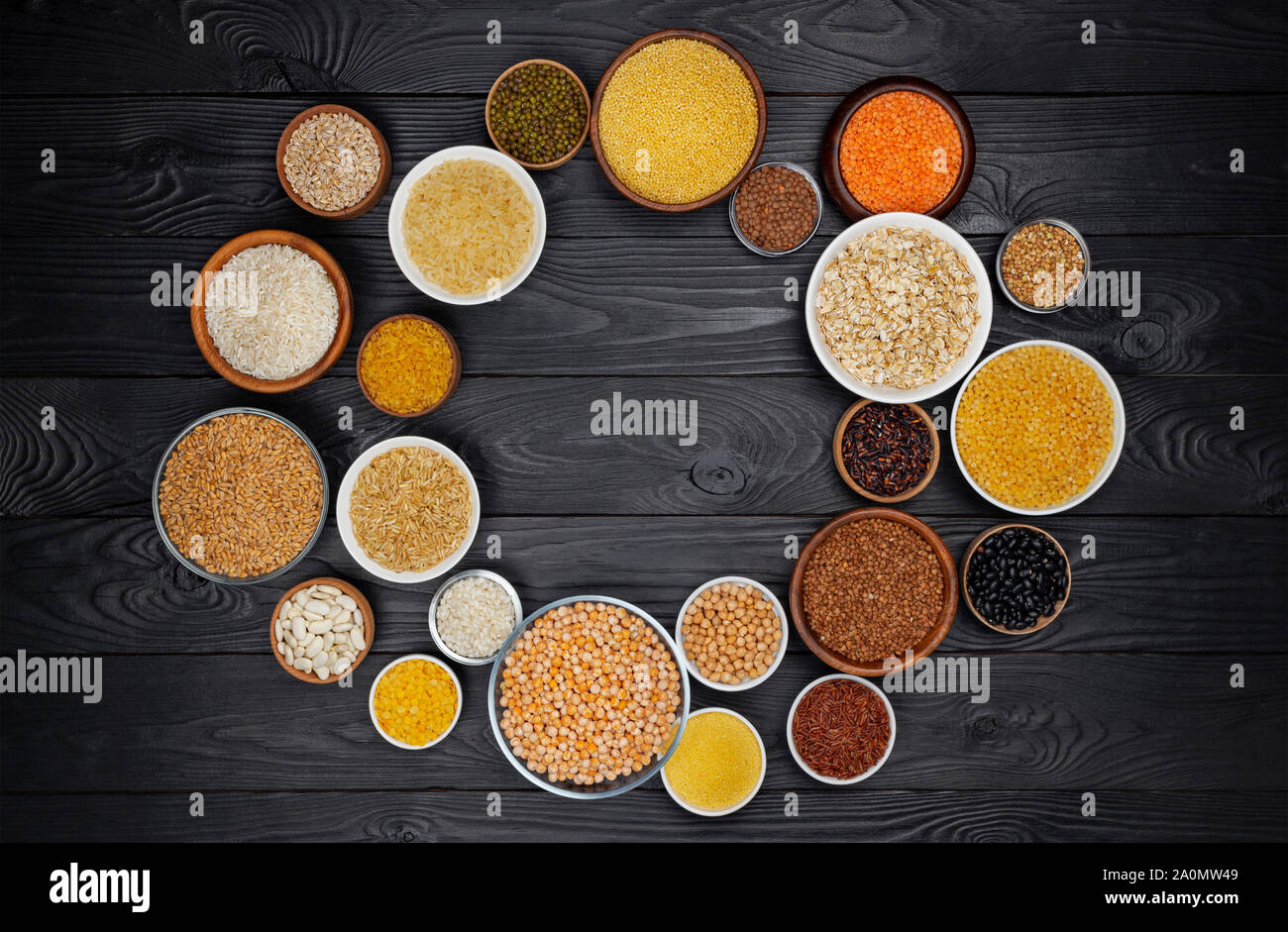 Cornice rotonda di cereali, granaglie, semi, semole, legumi e fagioli in ciotole, dieta ad alto contenuto di fibre concetto, vista dall'alto di porridge raccolta su legno nero ba Foto Stock