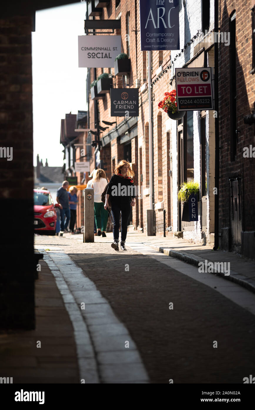 Una donna di età (30s) cammina verso la risoluta verso Leen Lane, un piccolo vicolo messo in ombra da edifici in centro di Chester. Un soleggiato aperto area dietro. Foto Stock Una donna di età (30s) cammina verso la risoluta verso Leen Lane, un piccolo vicolo messo in ombra da edifici in centro di Chester. Un soleggiato aperto area dietro. Foto Stock