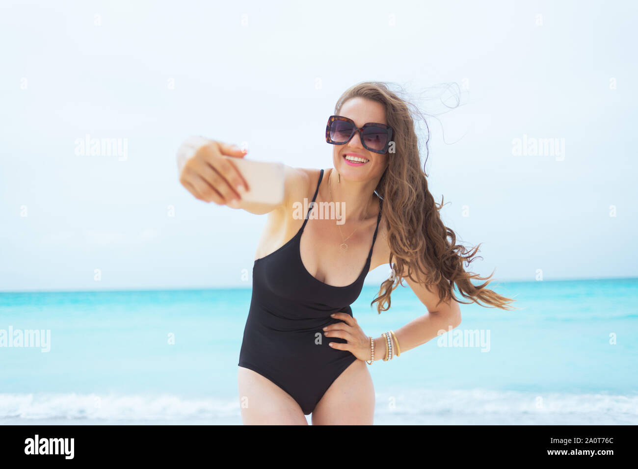 Sorridente giovane età media della donna in occhiali da sole prendendo selfie con telefono su una spiaggia di sabbia bianca. Foto Stock