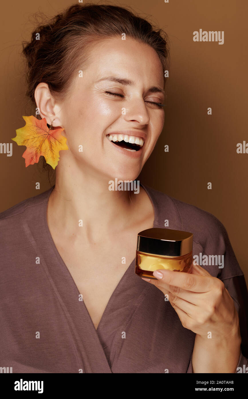 Ciao autunno. Ritratto di sorridere moderno medio evo woman in un accappatoio con foglie di autunno orecchini azienda crema per il viso contro lo sfondo di bronzo. Foto Stock