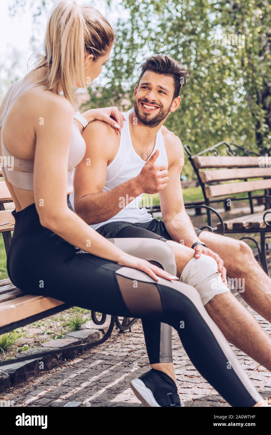 Feriti sportivo sorridente e mostrando un pollice alzato durante la seduta sul banco vicino sportive Foto Stock