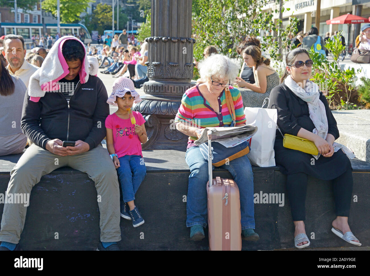 La gente seduta su una parete. Gruppo misto. Uomo al telefono, bambino, old Lady & un'altra. Foto Stock