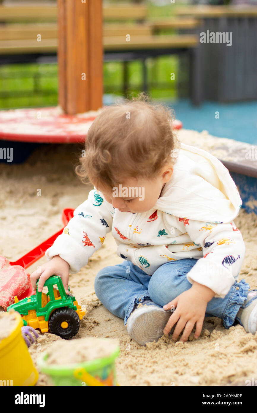 Baby girl boy in una felpa con cappuccio bianco e jeans gioca con un  trattore giocattolo sul parco giochi all'aperto. Felice Caucasian bambino 1  anno di età in una passeggiata, por verticale