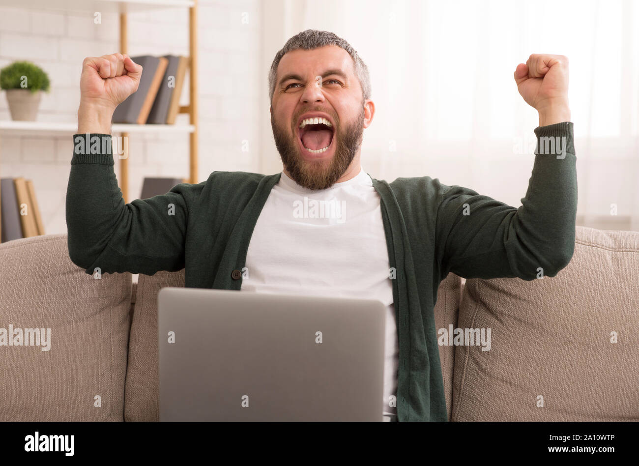 Eccitato uomo barbuto celebra il successo nella parte anteriore del computer portatile Foto Stock