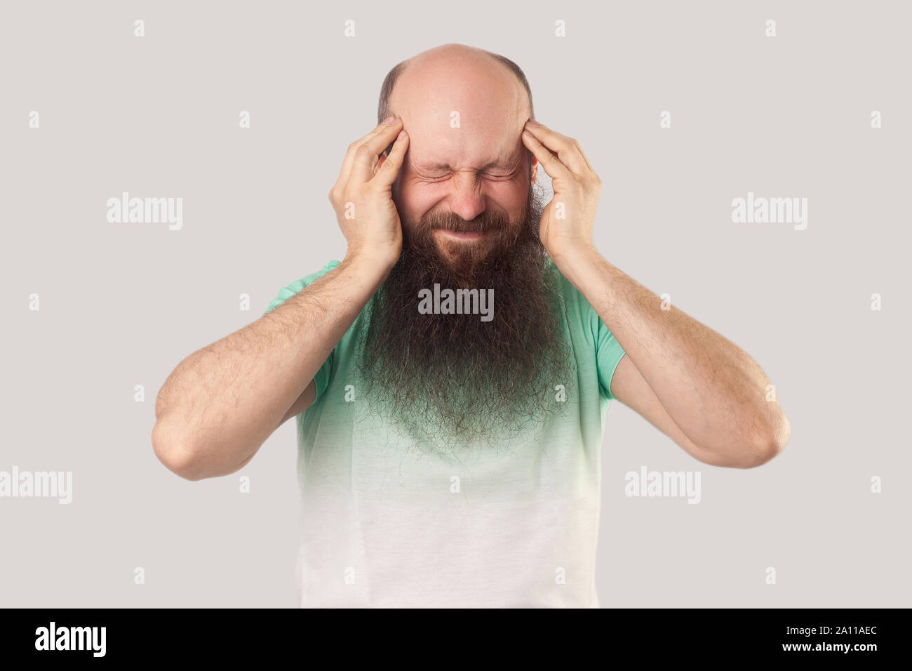Mal di testa, pensando o confusione. Ritratto di mezza età uomo calvo con barba lunga in verde chiaro t-shirt e permanente tenendo la sua testa doloroso. interna Foto Stock