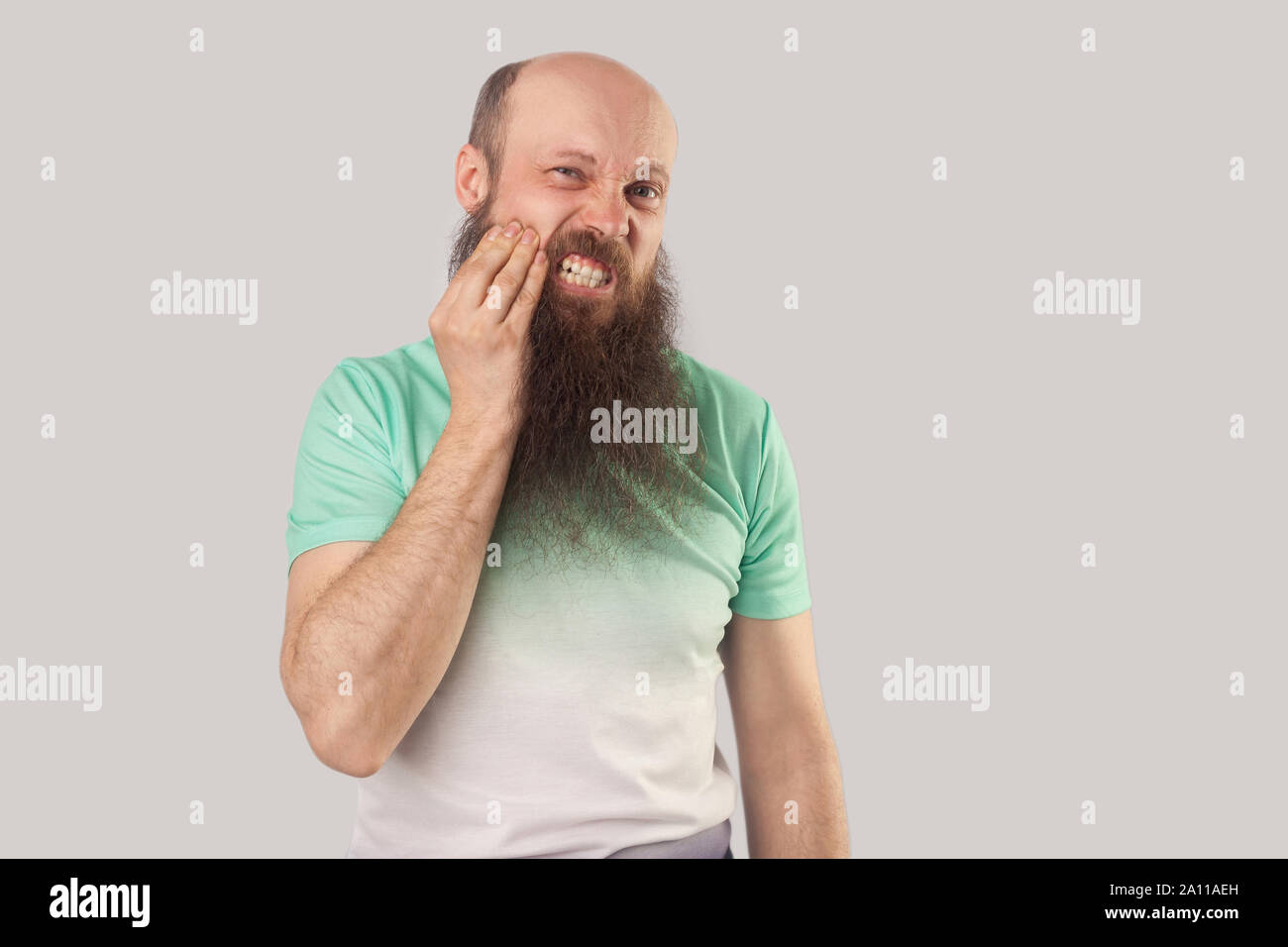 Il mal di denti. Ritratto di malati di mezza età uomo calvo con barba lunga in verde chiaro t-shirt in piedi toccano la guancia perché sentire dolore su i suoi denti. Foto Stock