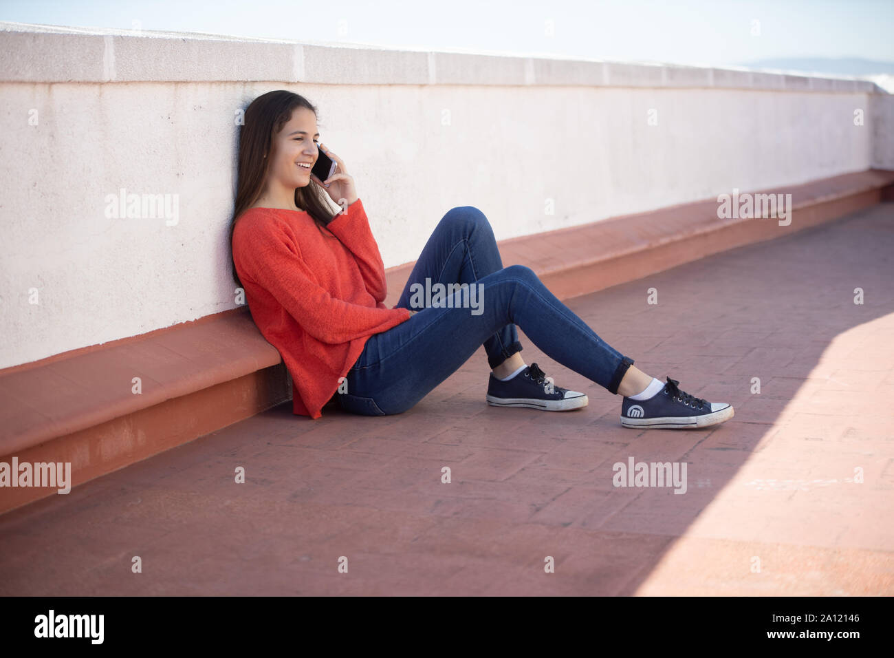 Chica adolescente feliz hablando por teléfono móvil sentada en la azotea Foto Stock
