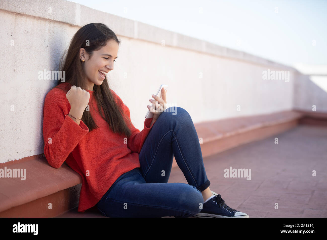 Chica adolescente celebrando buenas noticias on line sentada en la azotea Foto Stock