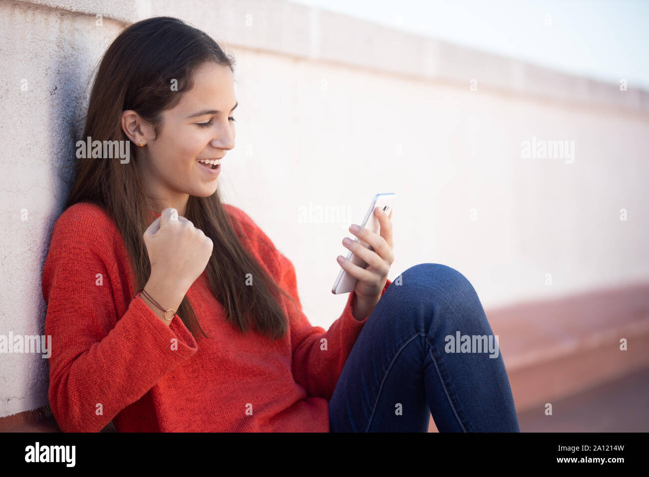 Chica adolescente celebrando buenas noticias on line sentada en la azotea Foto Stock