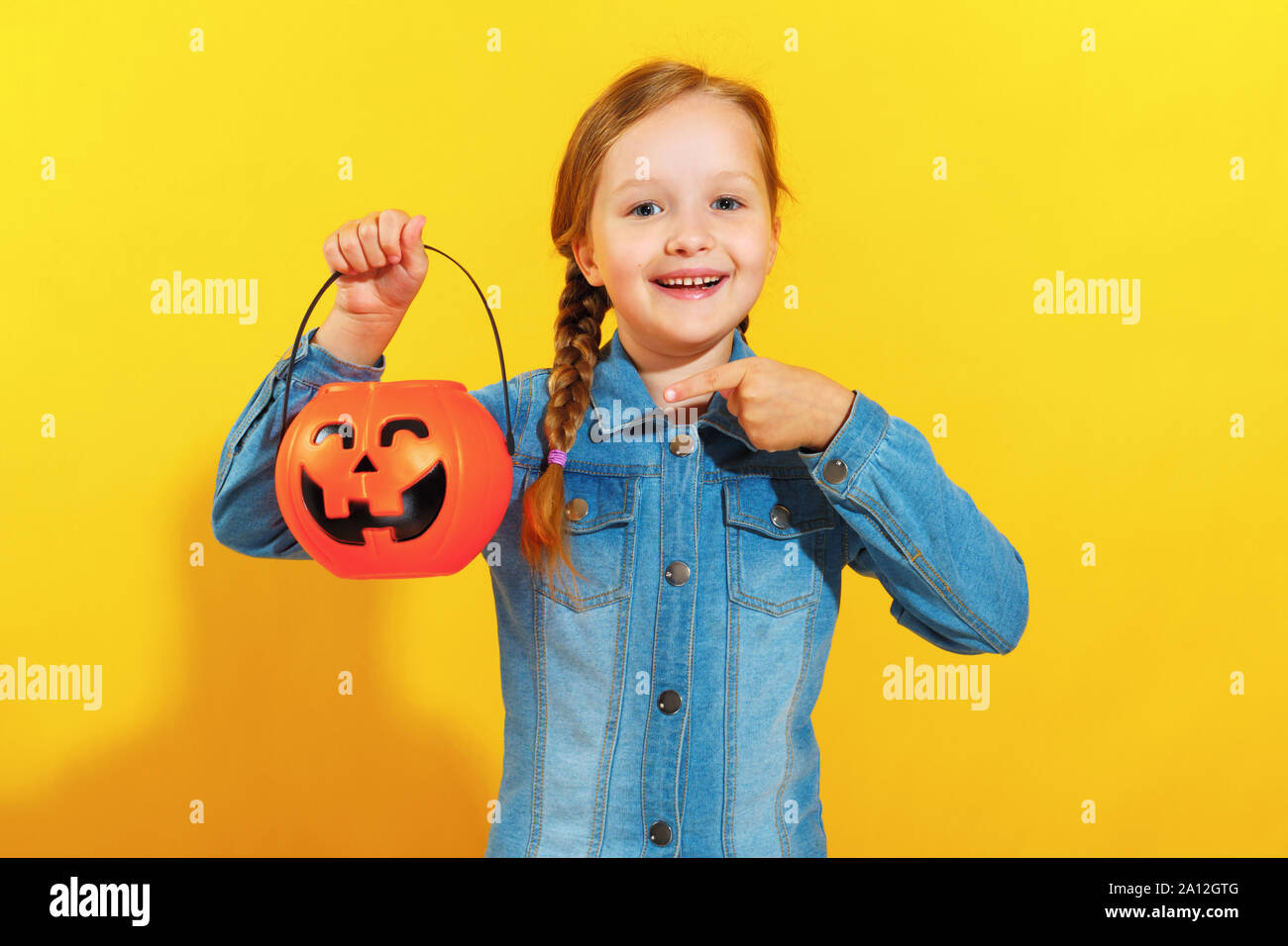 Halloween. Bambina in una camicia di denim su sfondo giallo. Il bambino mostra un dito su una benna di zucca lanterna di jack. Foto Stock