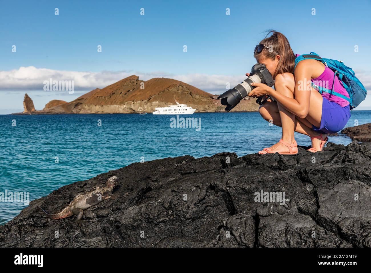 Galapagos fotografare turistica Iguana marina sull'isola di Santiago in Isole Galapagos. La nave di crociera e il pinnacolo di roccia e Bartolome isola in background. Galapagos famosa nave da crociera tour destinazione. Foto Stock