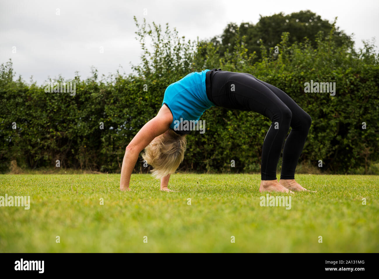 Una donna di mezza età a praticare yoga a piedi nudi fuori in un parco erbosa. Indossa una brillante giubbotto blu e nero leggings. Lo stile di yoga è lei Foto Stock