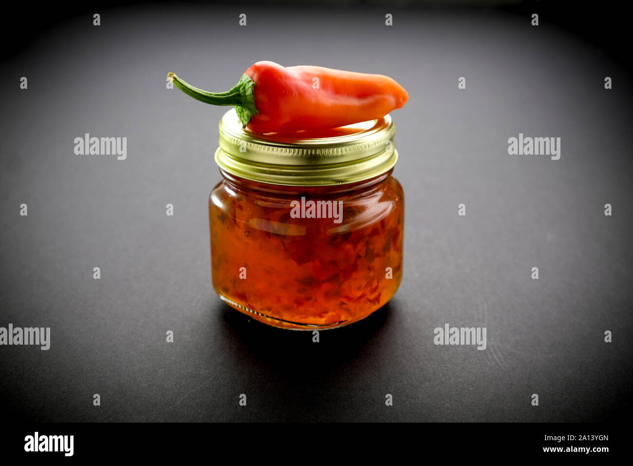 Piccolo vasetto in vetro di jalapeño jelly illuminato dalla luce dalla parte posteriore e laterale con una banana rosso pepe sulla parte superiore del barattolo Foto Stock